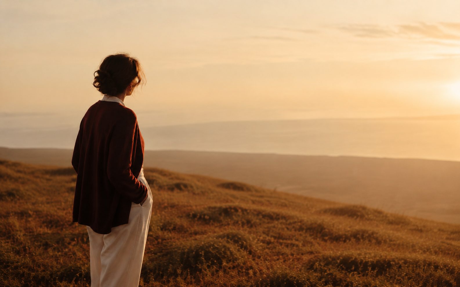 A woman standing alone on a hillside at sunset, looking out over the horizon
